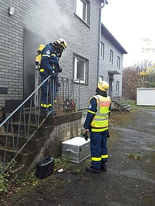 Ein Helfer mit angelegtem Atemschutzgerät steht vor einer Haustür, aus der Rauch dringt. Ein Ausbilder spricht mit dem Helfer.